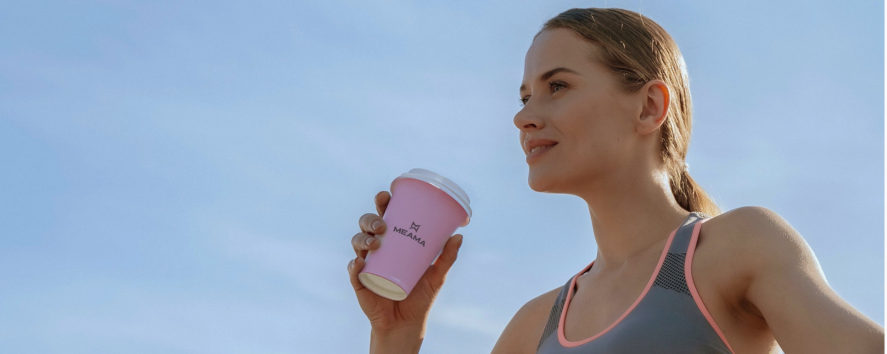 Eine Frau im sportlichen Outfit hält einen Meama Becher in der Hand hinter ihr ist nur blauer Himmel zu sehen.