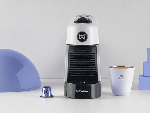 Die weiße Espresso Kapselmaschine steht mittig auf weißem Hintergrund, links davon eine Espresso Kapsel, rechts davon eine weiße Tasse mit Meama Logo