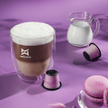 Ein Kaffee mit flaumigen Milchschaum steht neben einem Teller mit Macrons, Vanille Espresso Kapseln liegen auf dem Tisch, dahinter ein Krug mit Milch darin