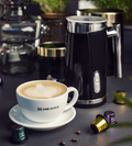 Der Schwarze Meama Milchschäumer steht in stimmungsvoller Kulisse, davor ein Cappuccino mit perfektem Milchschaum in einer Meama Kaffeetasse